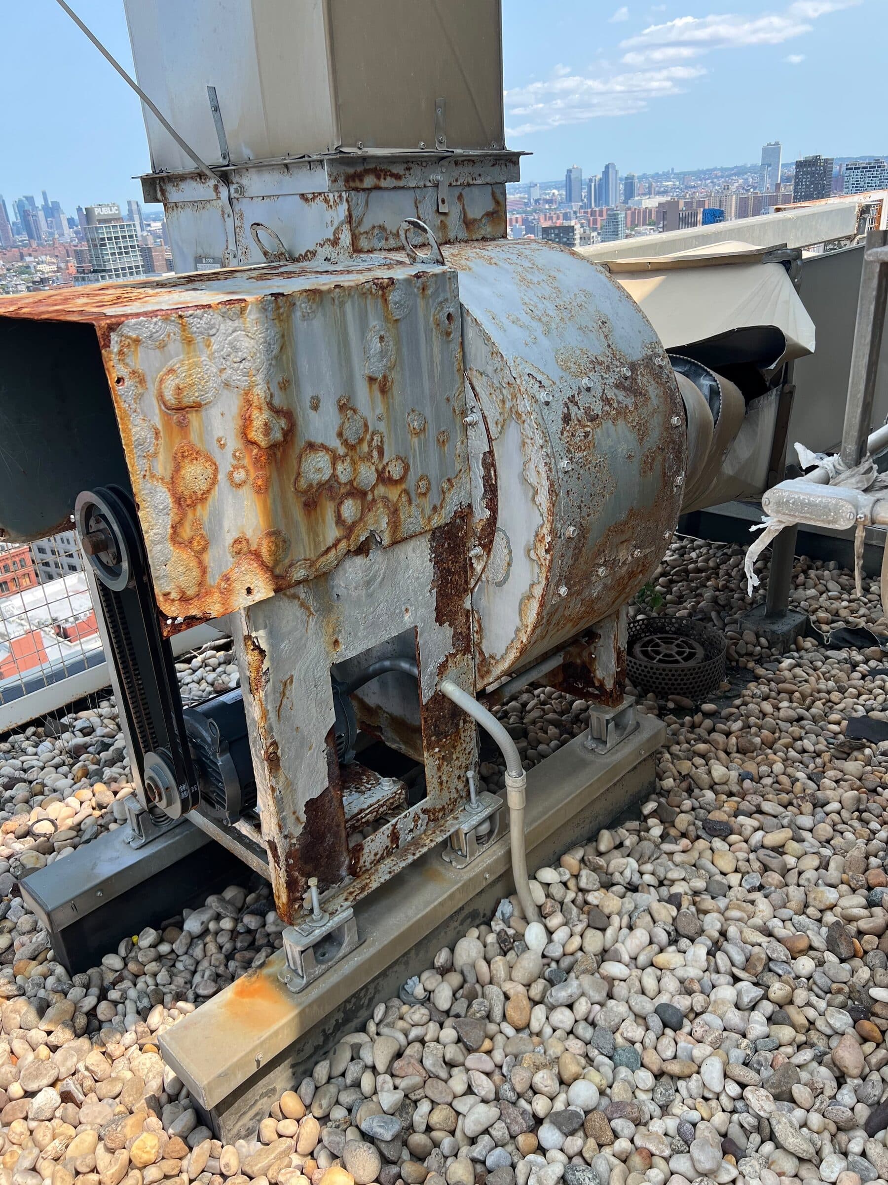 Heavily rusted Loren Cook belt-drive rooftop power ventilator on a downtown Manhattan roof with the city skyline behind, during a Vinco service visit