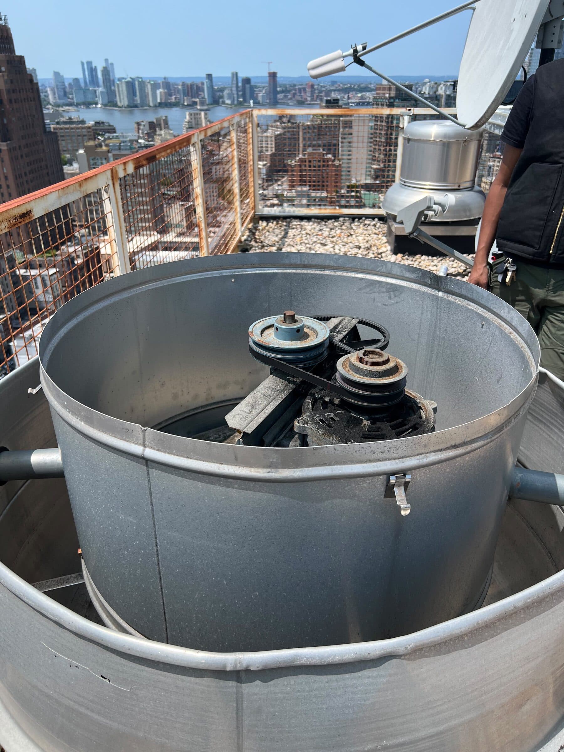 Open rooftop power ventilator during service, mushroom cap removed, belt and pulley drive visible inside the cylindrical housing, Hudson River skyline behind