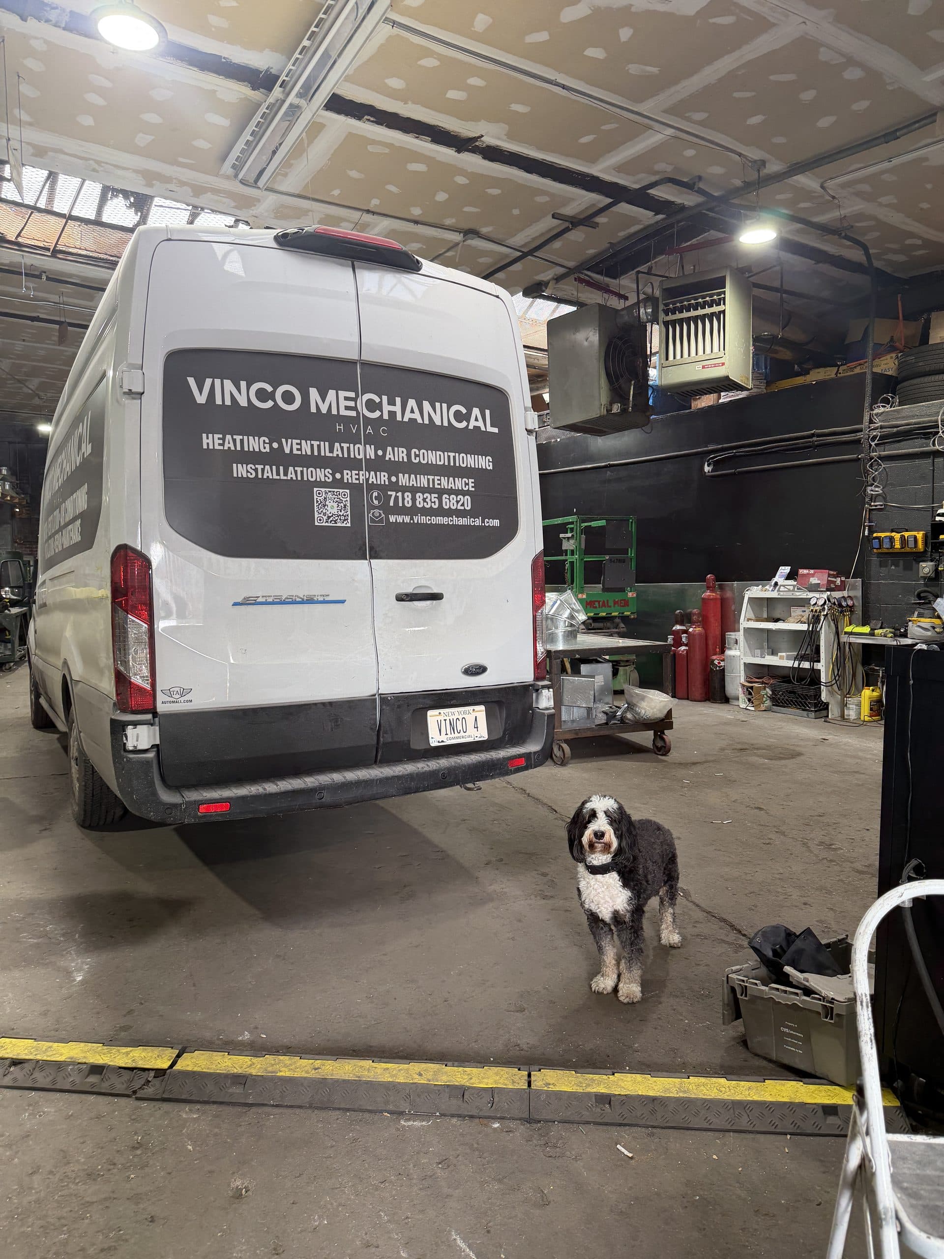 White Ford E-Transit electric van in Vinco Mechanical livery parked inside the 993 Grand Street shop, with the shop dog standing in front