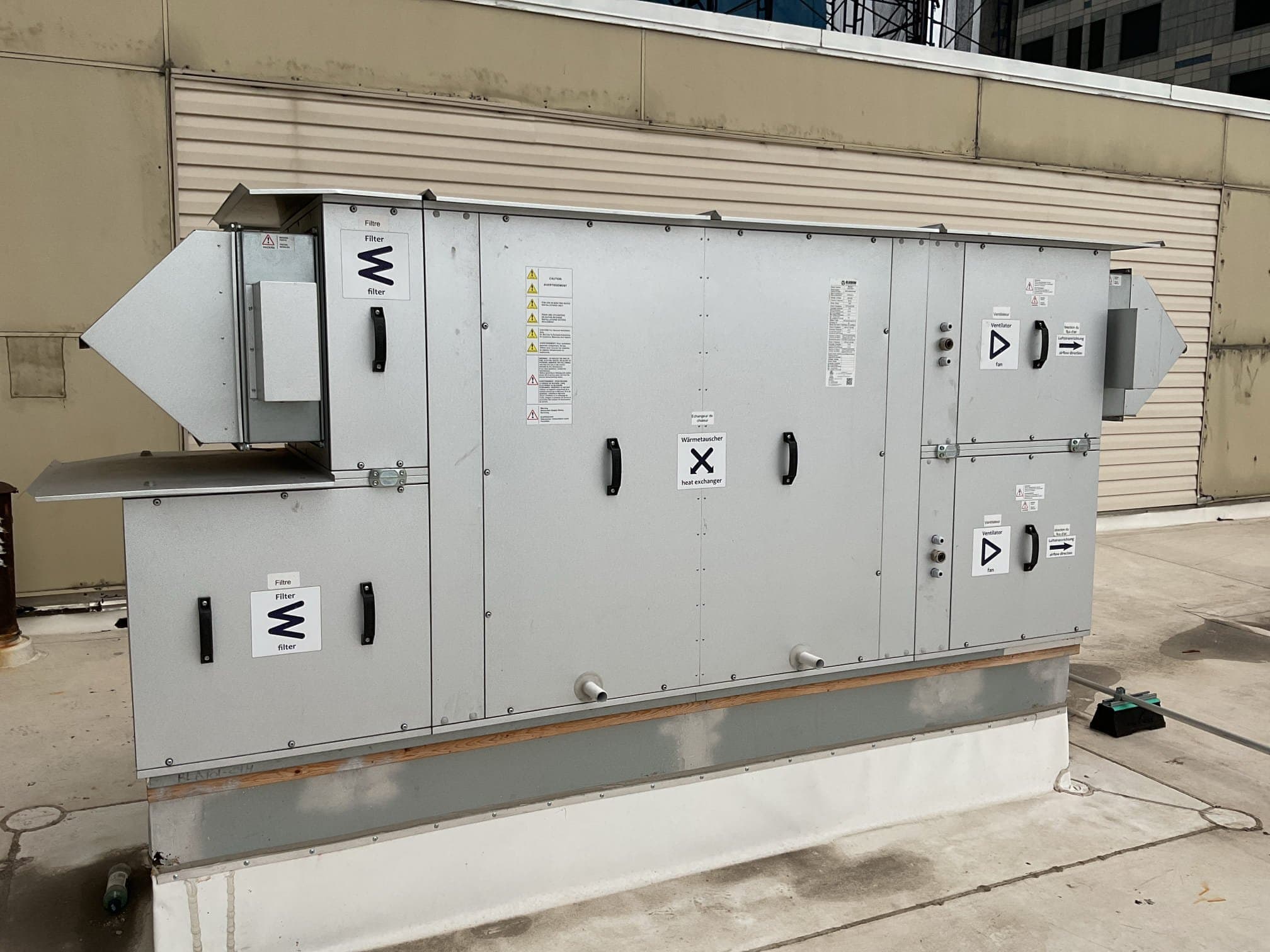Grey packaged rooftop air handler with filter and fan section labels, installed on a curb with an adjacent building behind