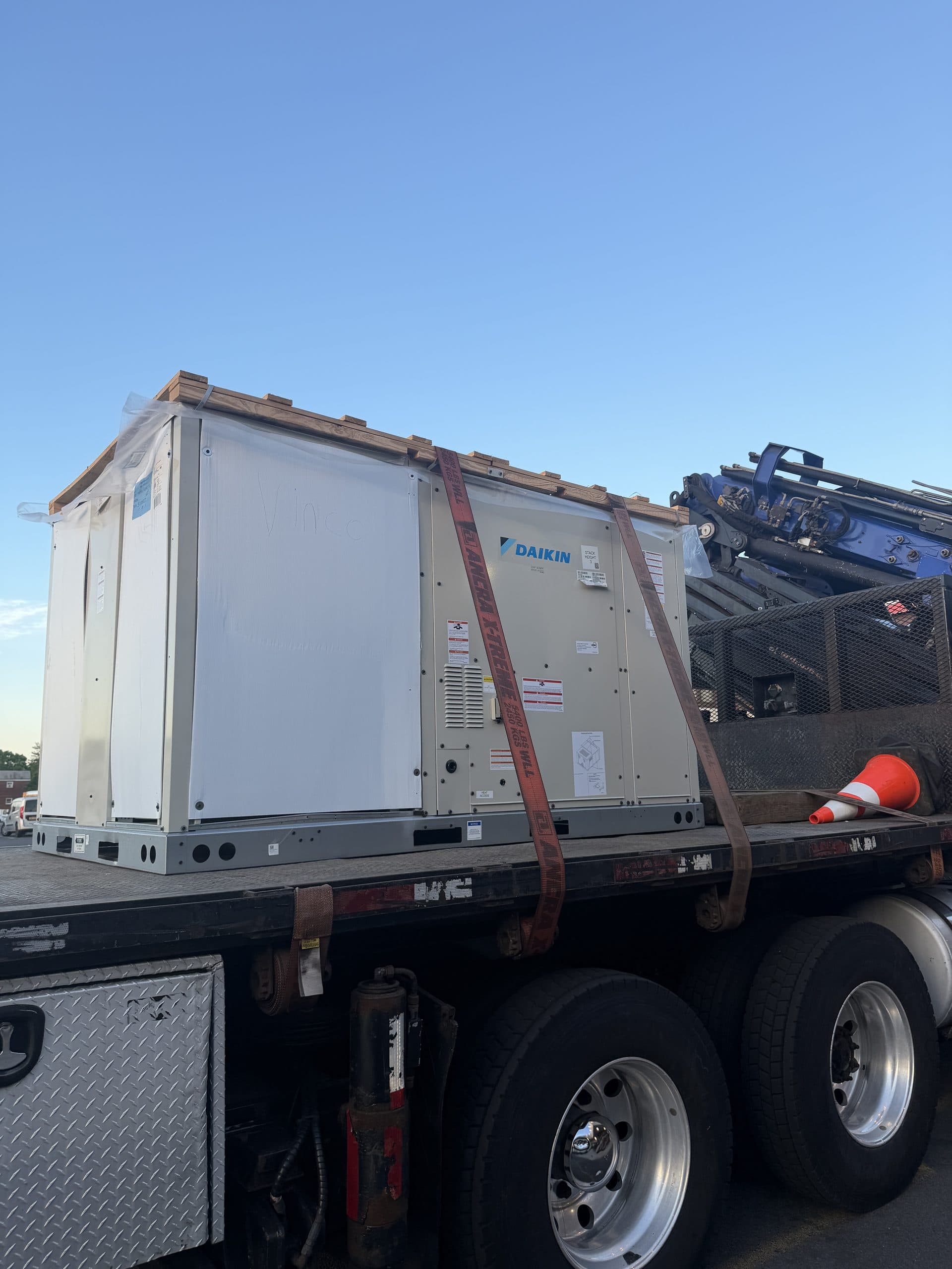 A crated Daikin rooftop unit strapped down on a Vinco Mechanical flatbed truck with a boom crane staged behind it