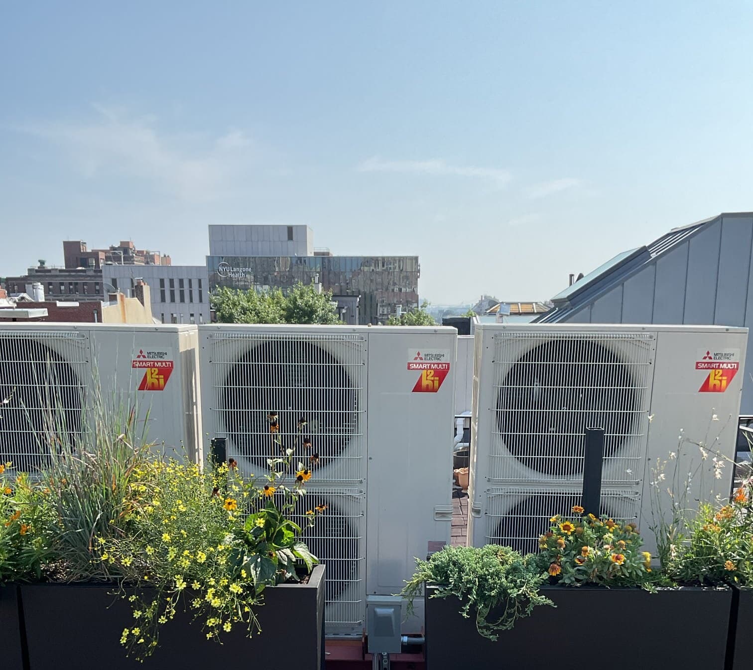 Three Mitsubishi Electric Smart Multi heat-pump condensers on a landscaped Brooklyn Heights roof deck with wildflower planters and the skyline behind
