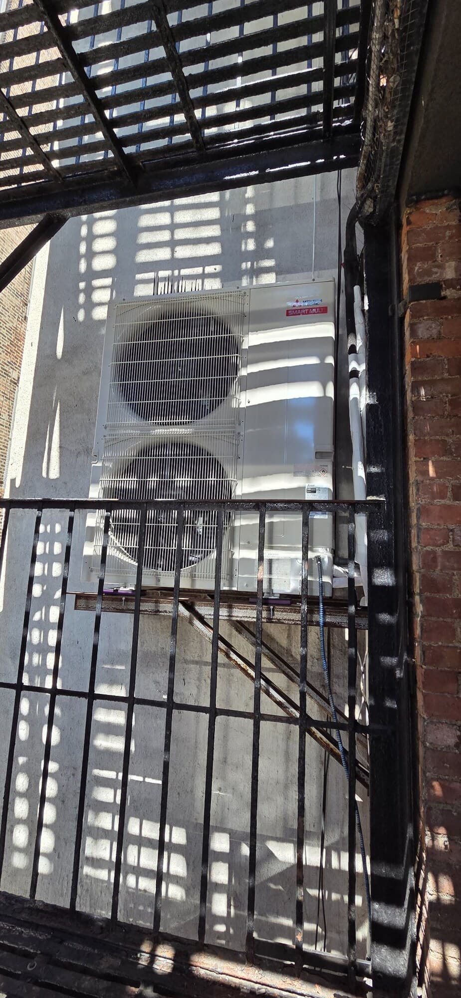 Mitsubishi R-454B multi-zone condenser mounted on a brick wall on Mercer Street in SoHo, seen through fire-escape railings