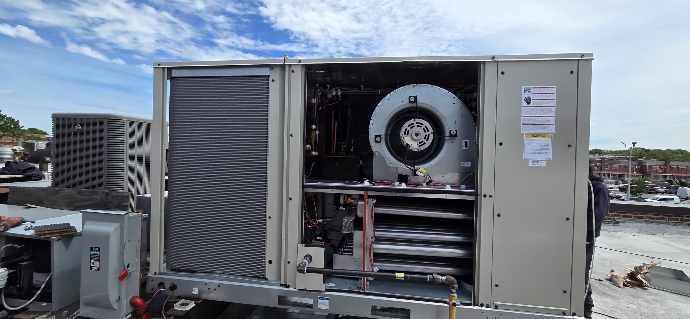 Rooftop packaged HVAC unit on a Brooklyn rooftop with the control-access panel open, showing the centrifugal blower, evaporator coil, and control board, a Rheem condenser on the adjacent curb