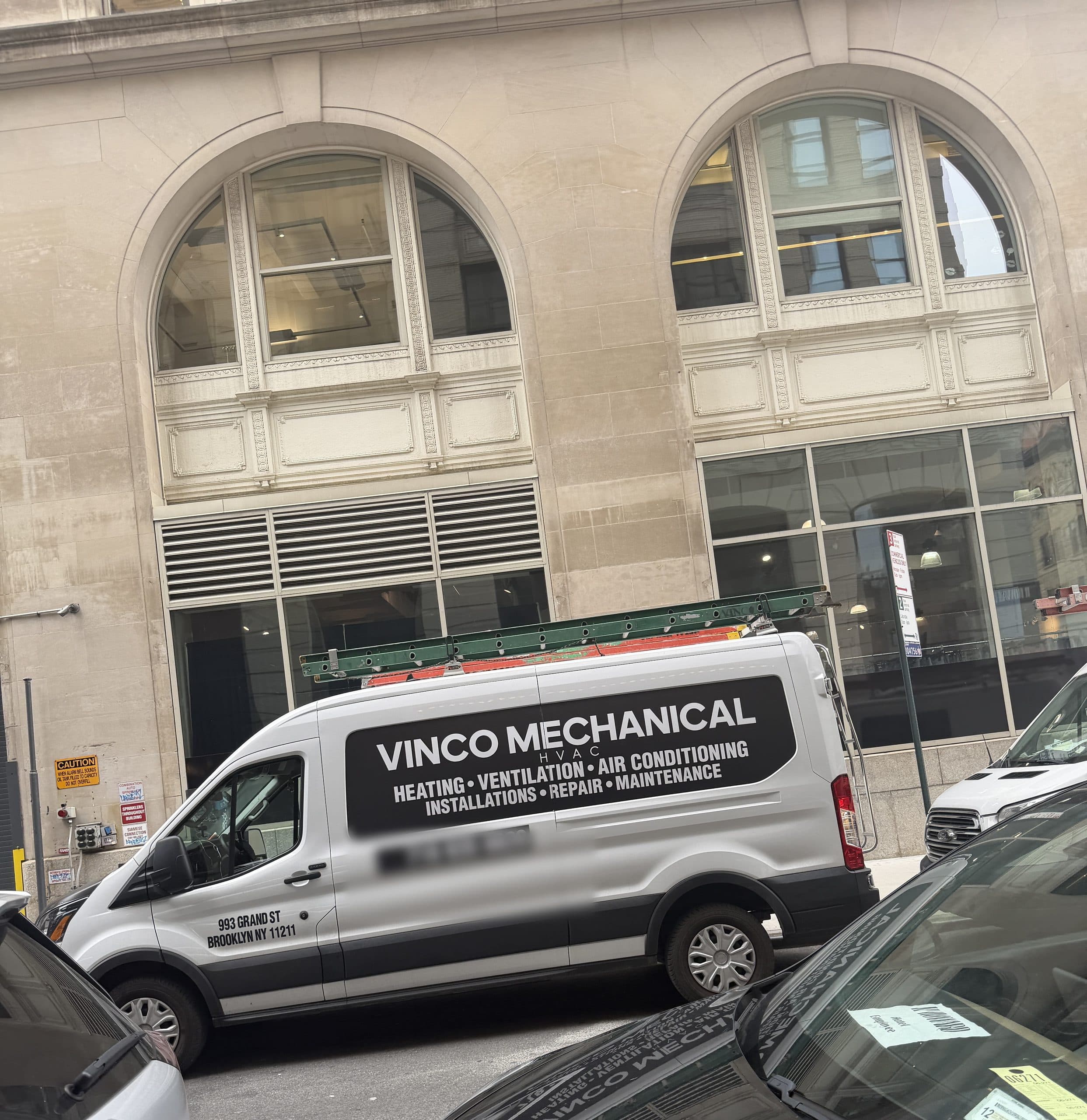 Vinco Mechanical shop van with ladders on the roof rack, parked at a job site outside a Manhattan building